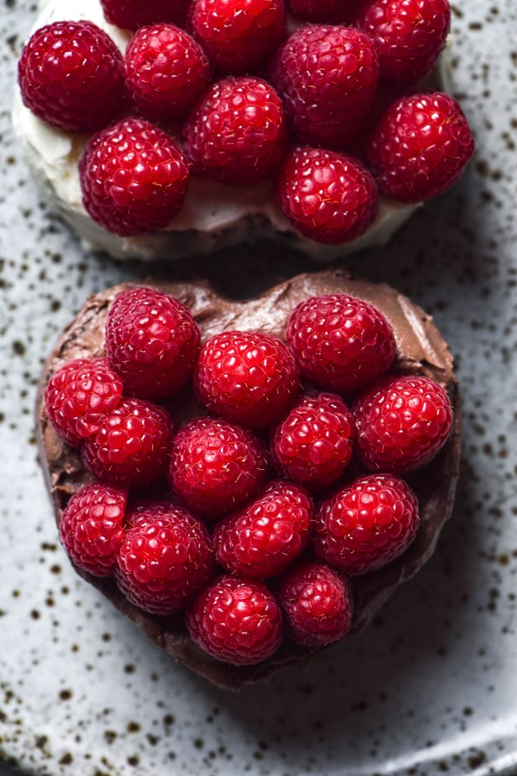 An aerial sunlit image two mini heart shaped gluten free chocolate cakes topped with raspberries