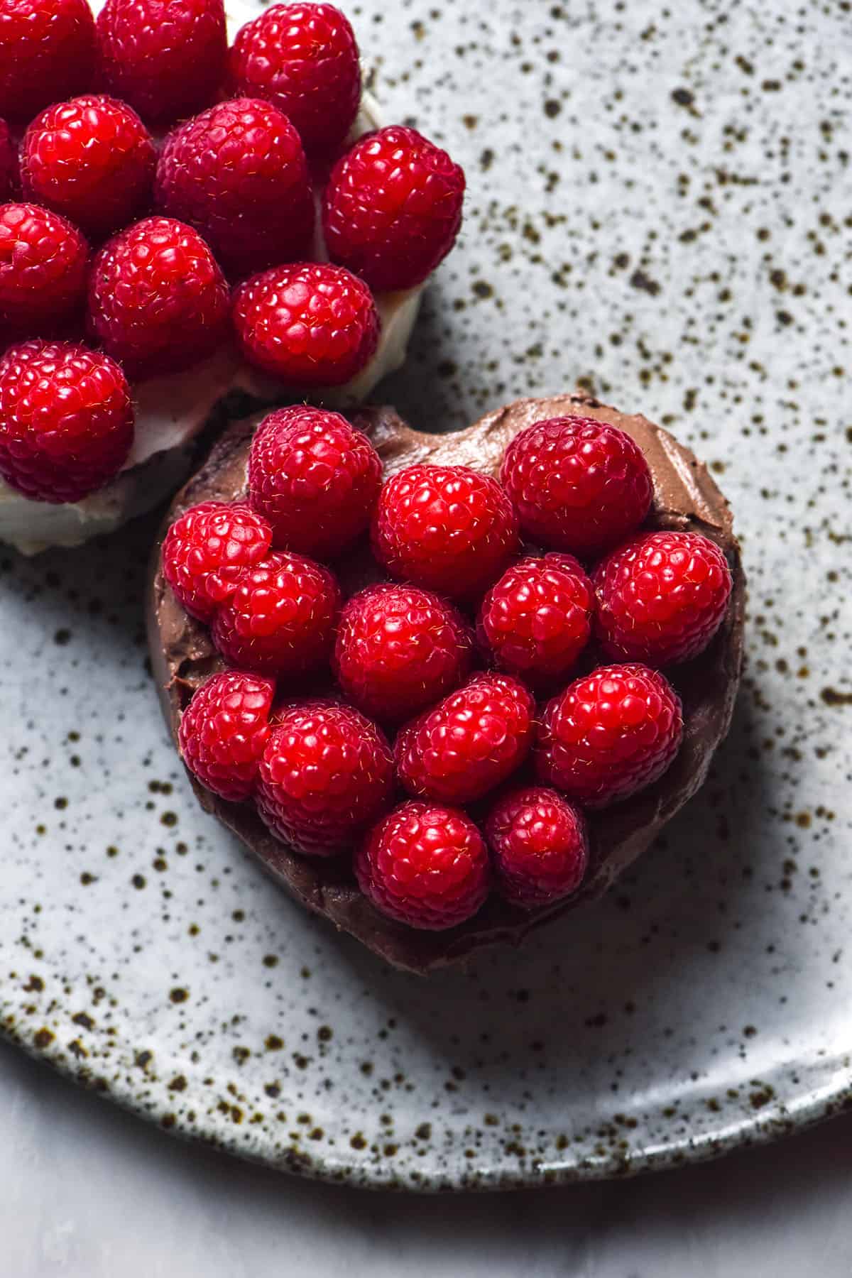 An aerial image of two mini chocolate cakes topped with raspberries. The cakes sit snugly on a white speckled ceramic plate atop a white marble table