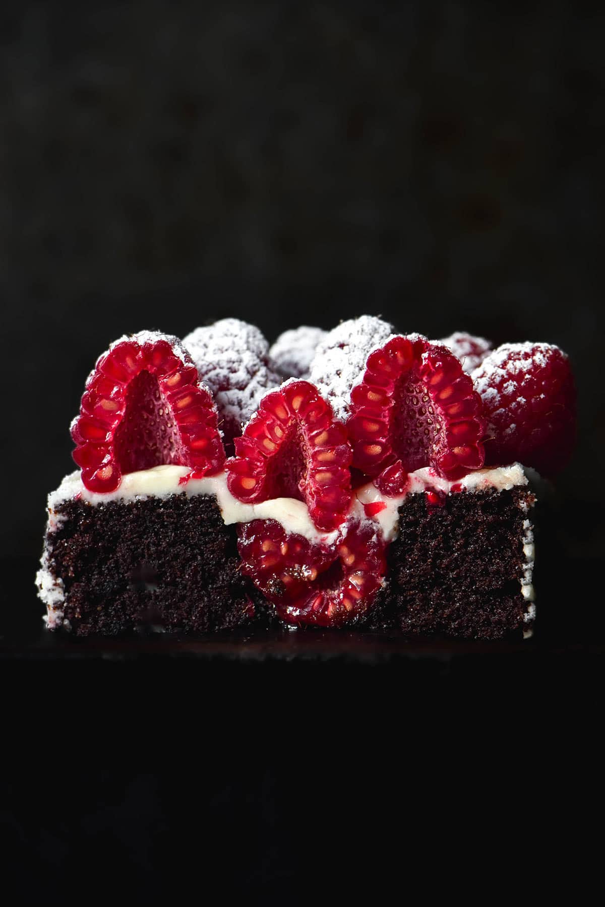 A dark and moody image of a mini gluten free chocolate cake that has been sliced down the centre, revealing a raspberry inside, vanilla icing and sliced raspberries on top