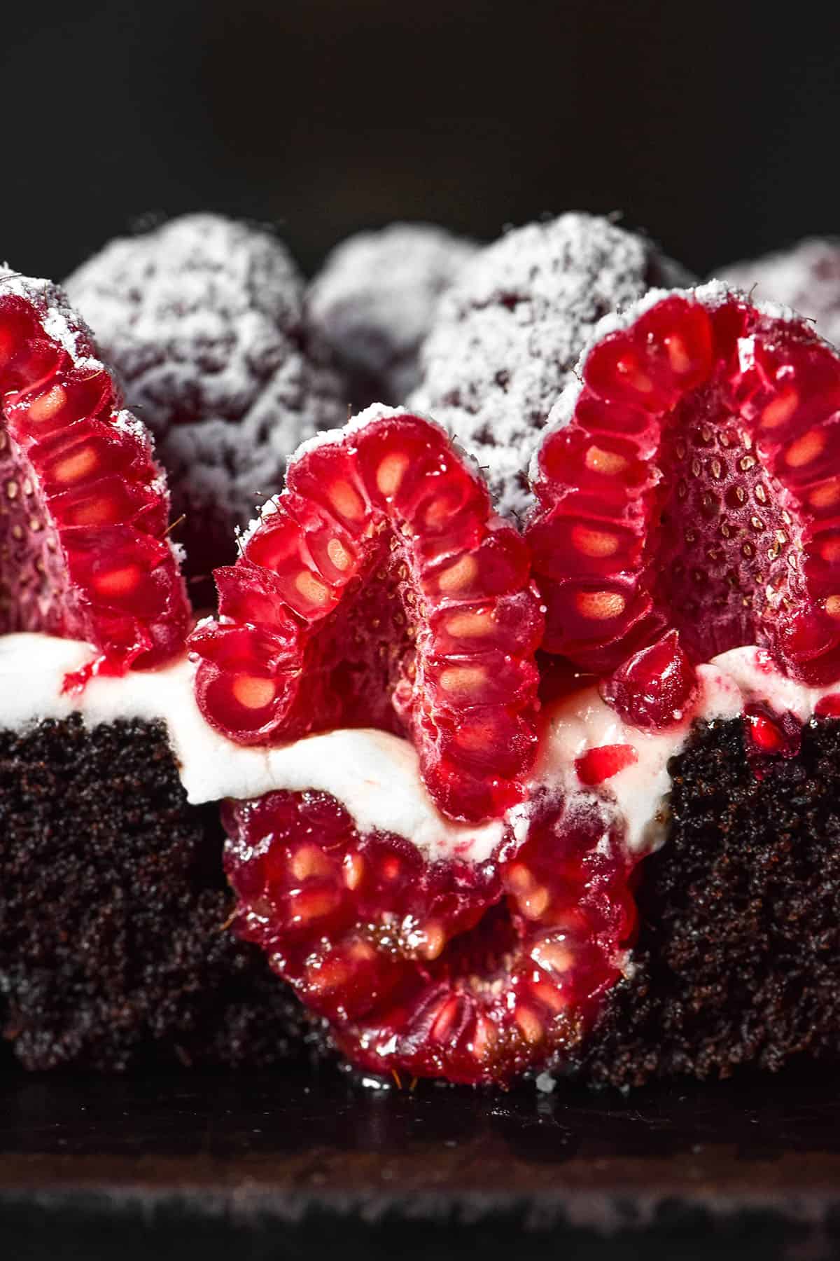 A macro close up image of a mini gluten free chocolate cake that has been cut in half, revealing a raspberry in the centre, vanilla icing and sliced raspberries on top