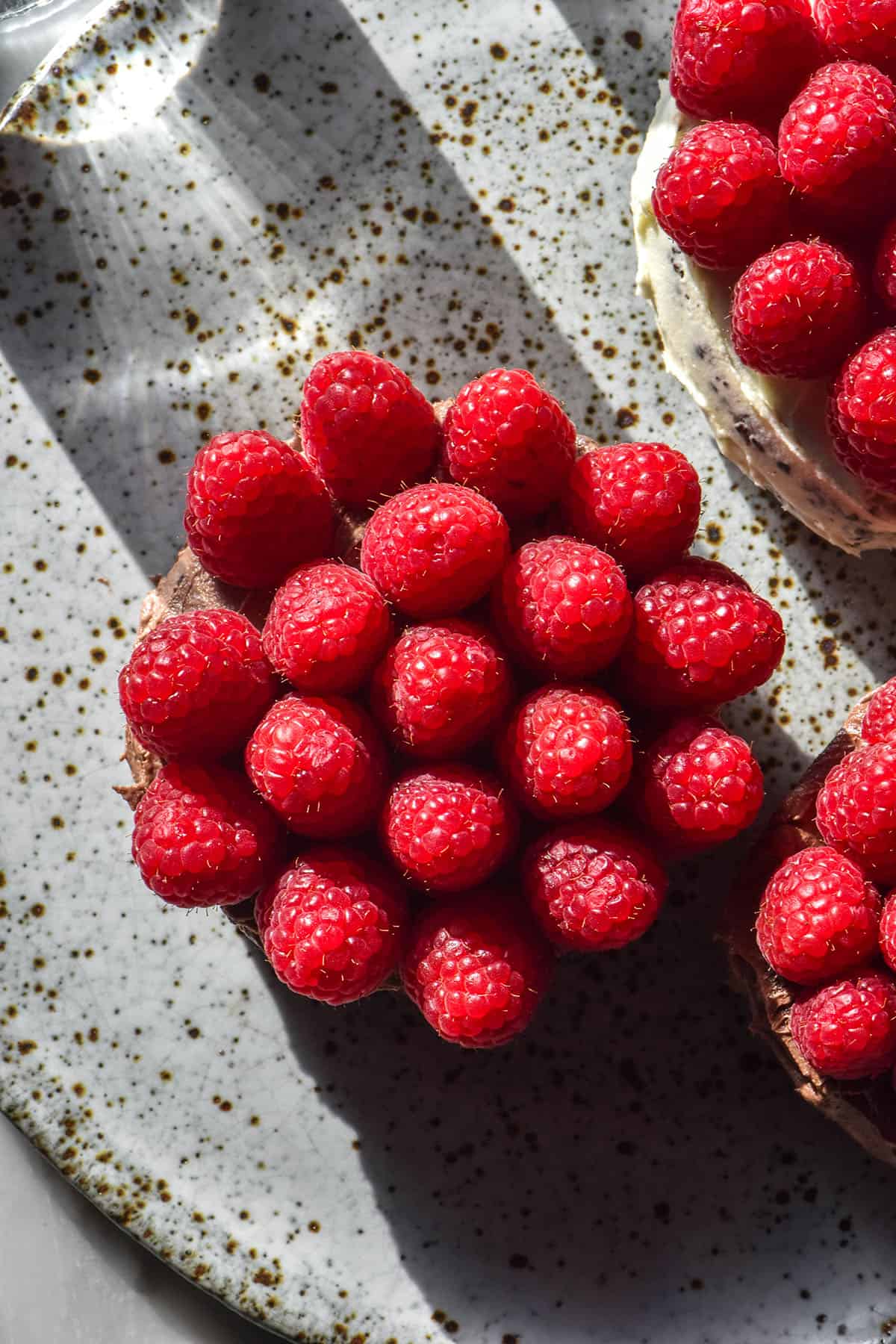 An aerial sunlit image of three mini gluten free chocolate cakes topped with raspberries on a white speckled ceramic plate atop a marble table 