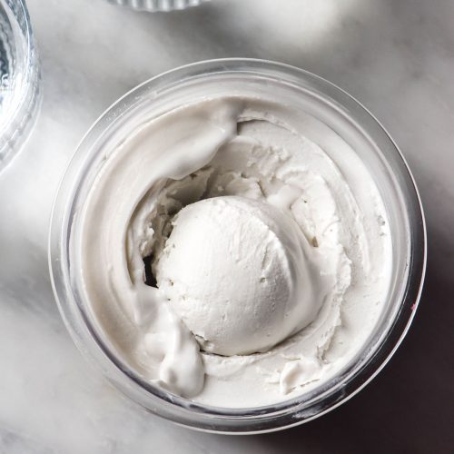 An aerial image of a tub of Ninja Creami coconut ice cream on a white marble table. Two sunlit glasses of water sit to the left of the tub.