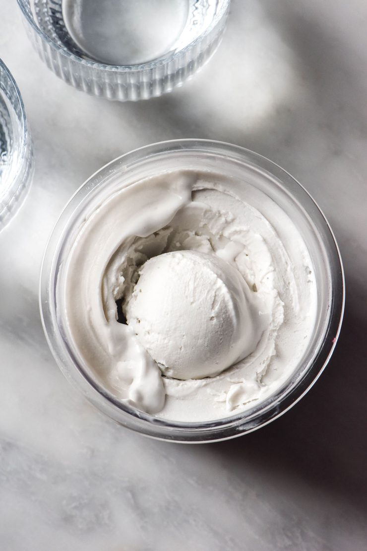 An aerial image of a tub of Ninja Creami coconut ice cream on a white marble table. Two sunlit glasses of water sit to the left of the tub.