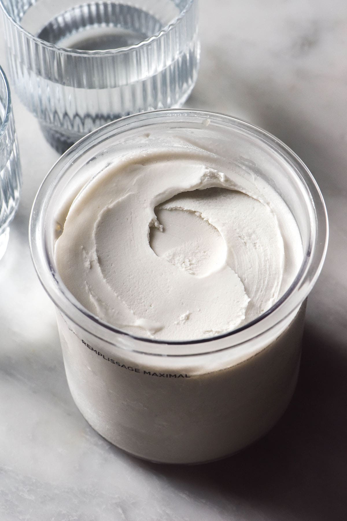 An aerial image of a tub of Ninja Creami coconut ice cream on a white marble table. Two sunlit glasses of water sit to the left of the tub.