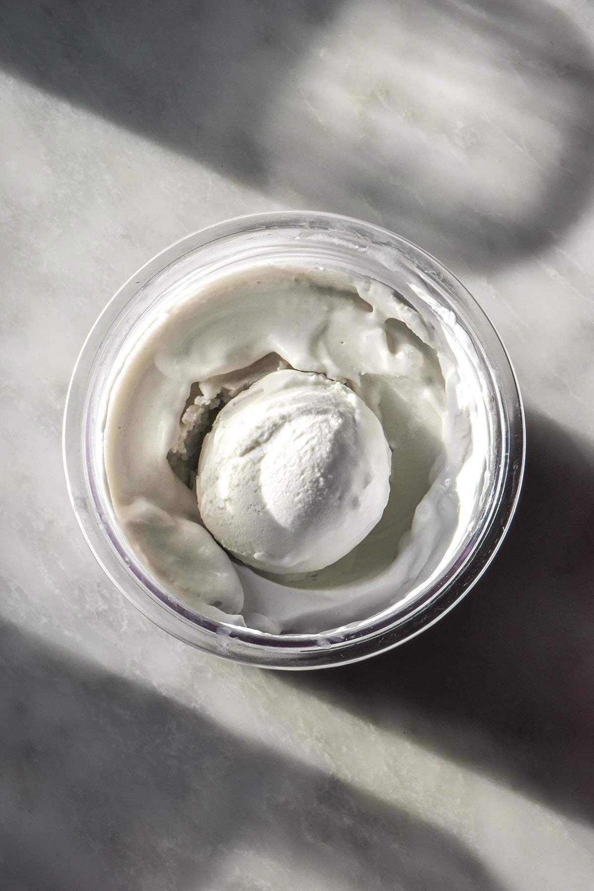 A sunlit aerial image of a tub of vegan coconut Ninja Creami ice cream on a white marble table. Two sunlit glasses of water sit to the left of the image and create a pattern across the marble. 