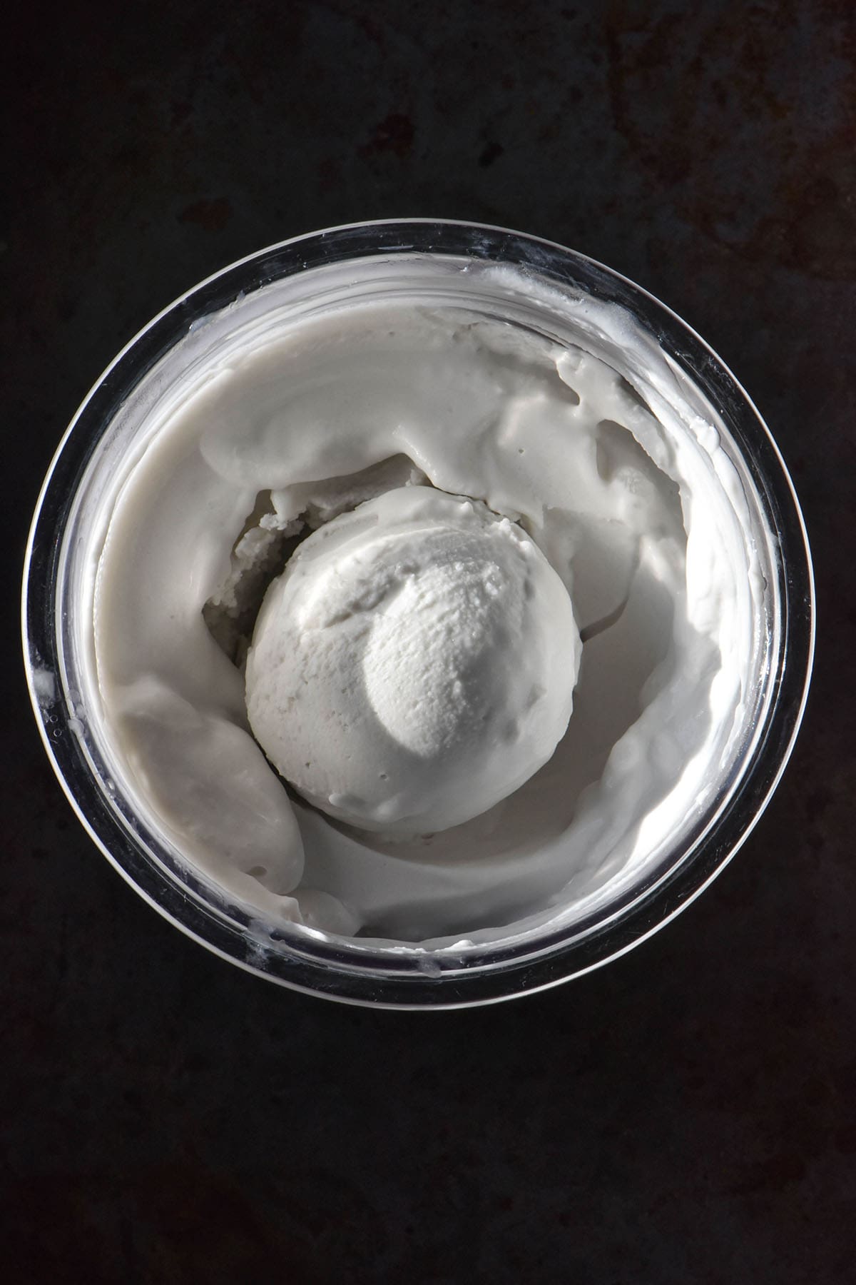 An aerial image of a tub of coconut Ninja Creami ice cream on a dark steel backdrop. The ice cream has a scoop casually resting in the centre.