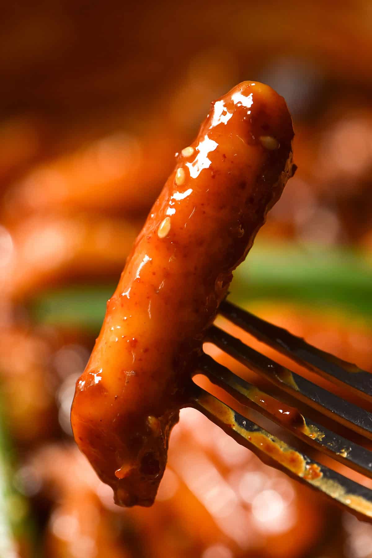 A close up macro image of a piece of vegan tteok on a fork set against a skillet filled with tteokbokki