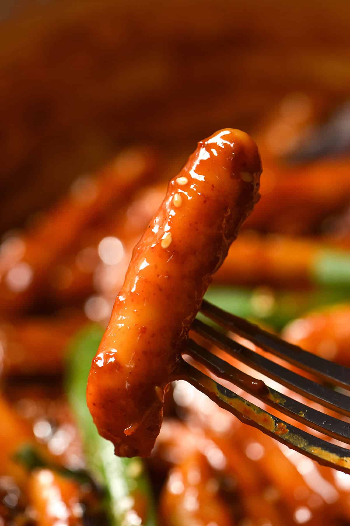 A close up macro image of a piece of vegan tteok on a fork set against the backdrop of a skillet filled with tteokbokki