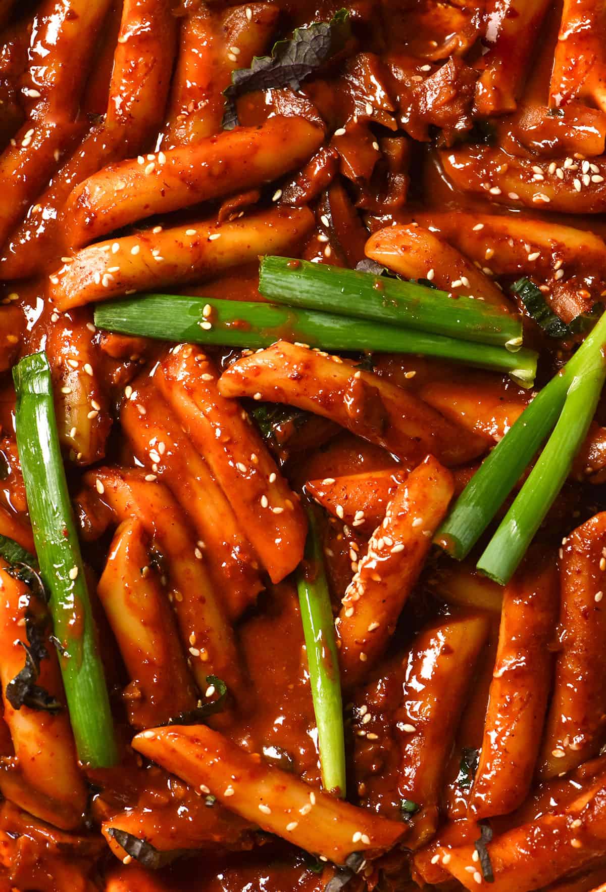 An aerial macro image of a skillet of vegan tteokbokki topped with spring onion greens, perilla leaves and sesame seeds