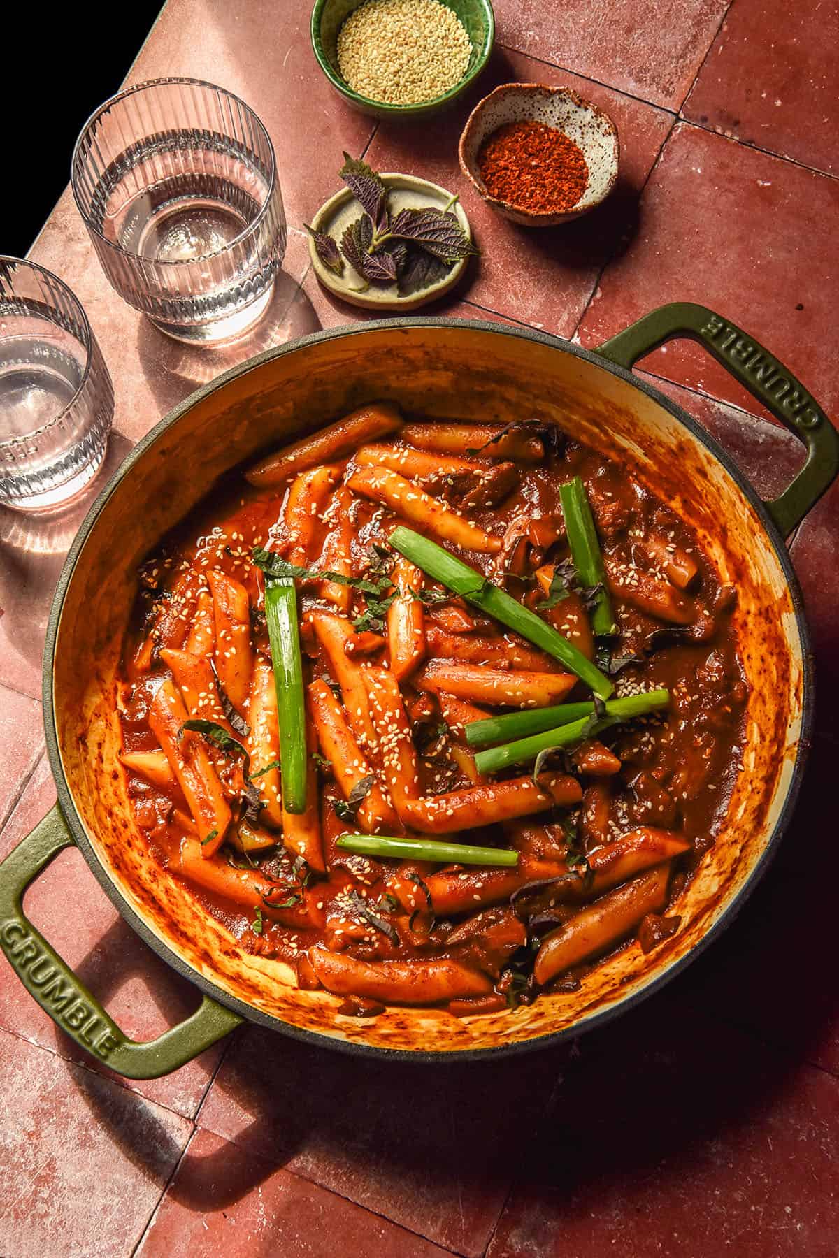 An aerial image of a skillet filled with vegan gluten free tteokbokki against a terracotta tile backdrop. Small bowls of ingredients and sunlit water glasses sit to the left of the skillet. 