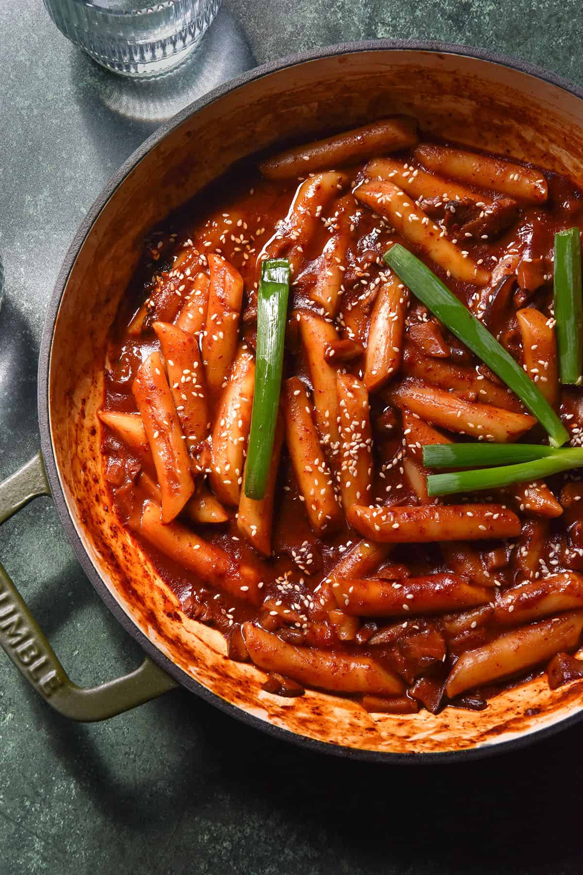 An aerial image of a green skillet filled with vegan gluten free tteokbokki on an olive green backdrop. Two glasses of water sit to the left of the skillet. 