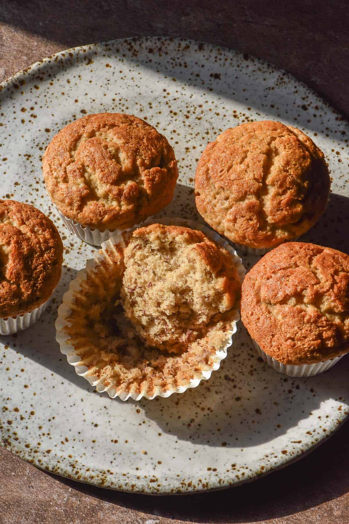 An aerial image of a fluffy gluten free vegan muffin on a white speckled ceramic plate atop a dark grey backdrop