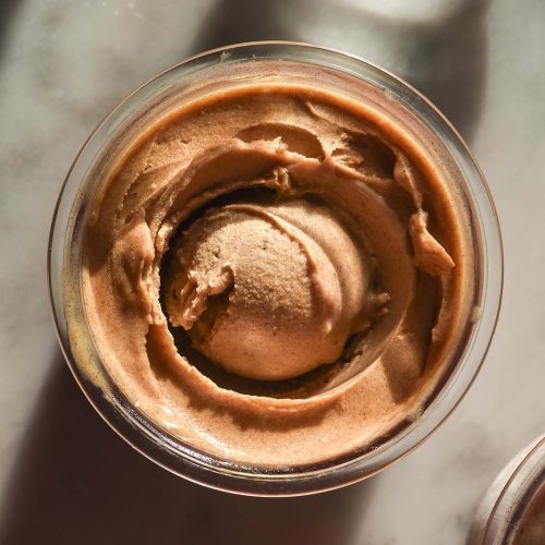 An aerial sunlit image of a tub of Ninja Creami sticky date protein ice cream on a white marble table. A glass of water sits to the top right of the image while a second tub of protein ice cream sits in the bottom left hand corner.