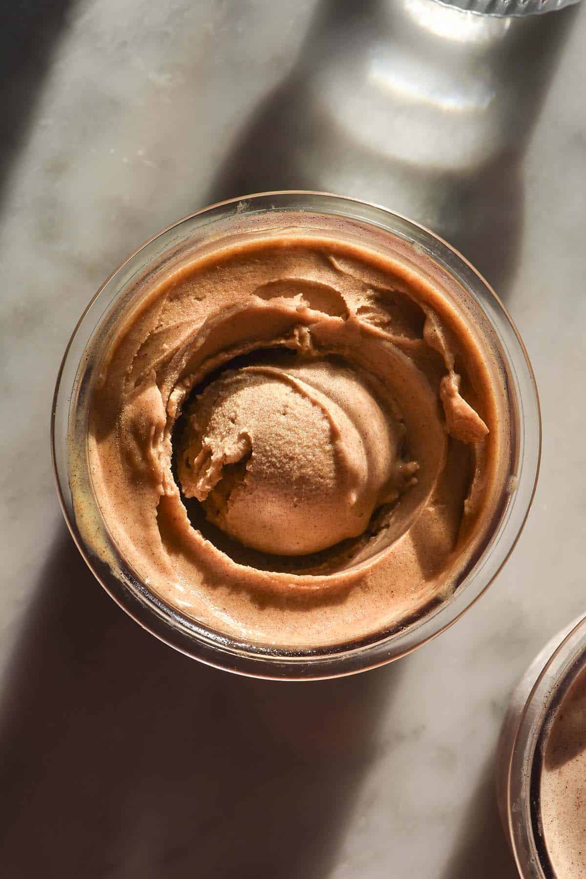 An aerial sunlit image of a tub of Ninja Creami sticky date protein ice cream on a white marble table. A glass of water sits to the top right of the image while a second tub of protein ice cream sits in the bottom left hand corner. 