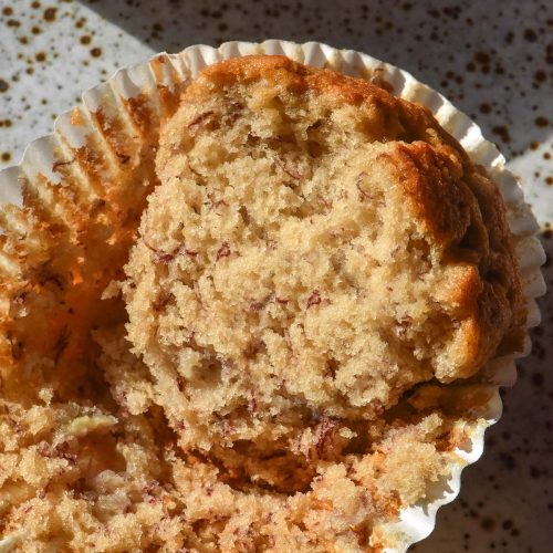 An aerial sunlit image of a gluten free banana muffin that has been cut in half, revealing the soft inner crumb. The muffin sits atop a white speckled ceramic plate in contrasting sunlight.