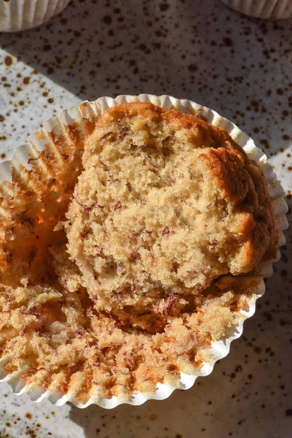 An aerial sunlit image of a gluten free banana muffin that has been cut in half, revealing the soft inner crumb. The muffin sits atop a white speckled ceramic plate in contrasting sunlight.