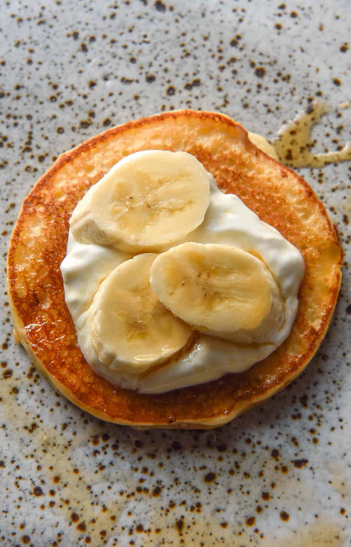 An aerial image of a gluten free cottage cheese pancake topped with maple syrup, yoghurt and sliced banana. It sits atop a white speckled ceramic plate.