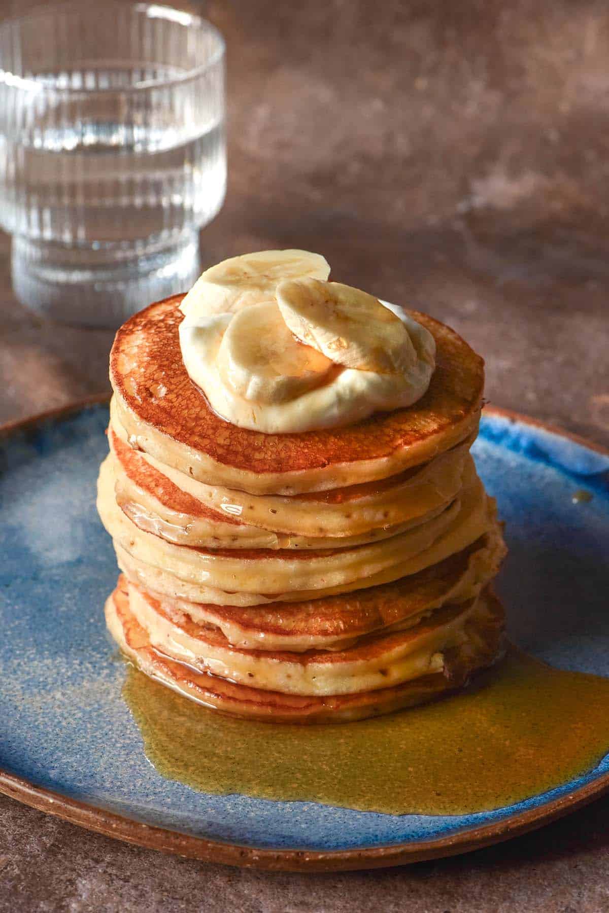 A side on image of a stack of gluten free cottage cheese pancakes on a bright blue ceramic plate against a dark backdrop. The pancakes have been topped with yoghurt, banana and a drizzle of maple syrup. 