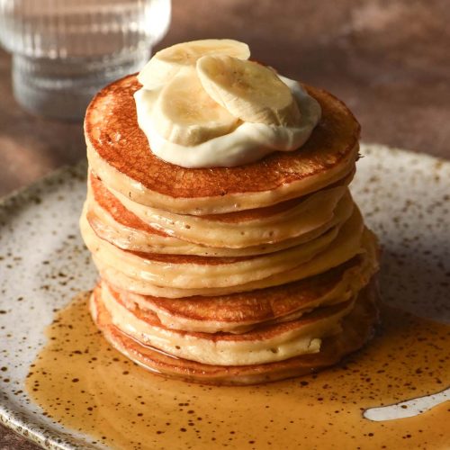 A side on image of a stack of gluten free cottage cheese pancakes on a white speckled ceramic plate against a dark brown backdrop. The pancakes have been drizzled with maple syrup and topped with a spoonful of yoghurt and sliced bananas.