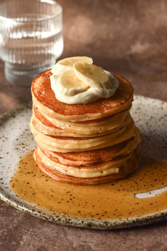 A side on image of a stack of gluten free cottage cheese pancakes on a white speckled ceramic plate against a dark brown backdrop. The pancakes have been drizzled with maple syrup and topped with a spoonful of yoghurt and sliced bananas.