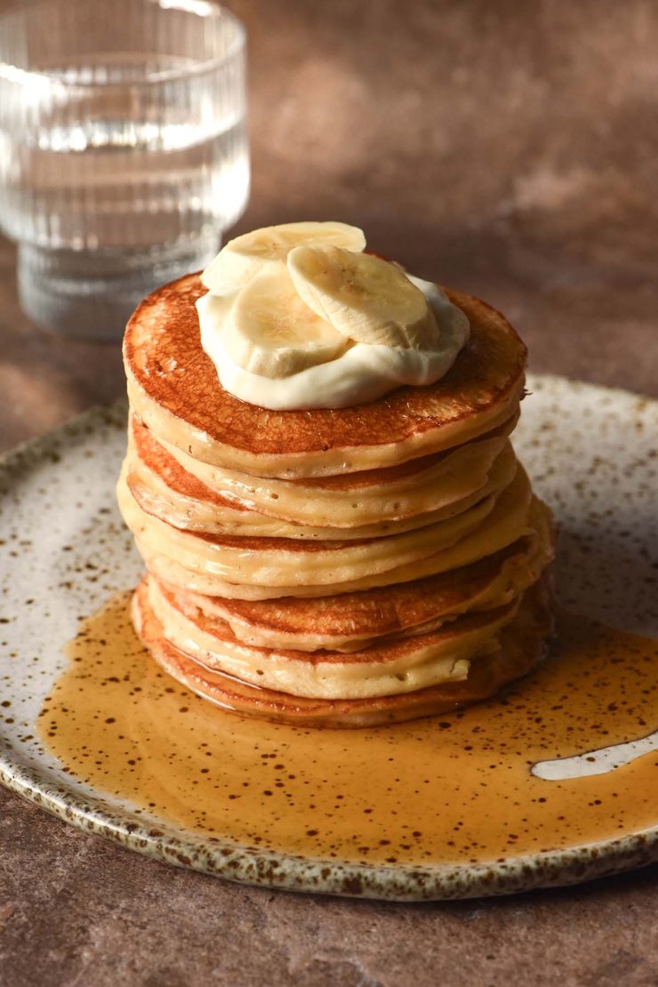 A side on image of a stack of gluten free cottage cheese pancakes on a white speckled ceramic plate against a dark brown backdrop. The pancakes have been drizzled with maple syrup and topped with a spoonful of yoghurt and sliced bananas.