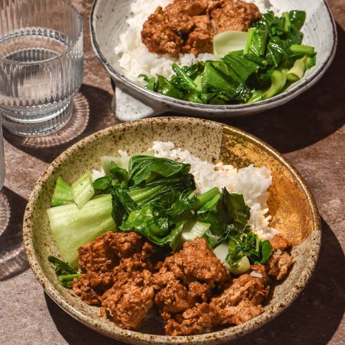 A side on image of two bowls filled with marinated tofu, rice and sautéed greens on a dark brown backdrop