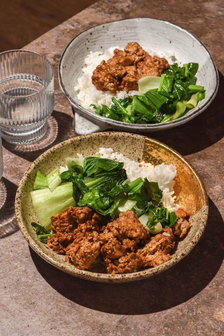A side on image of two bowls filled with marinated tofu, rice and sautéed greens on a dark brown backdrop