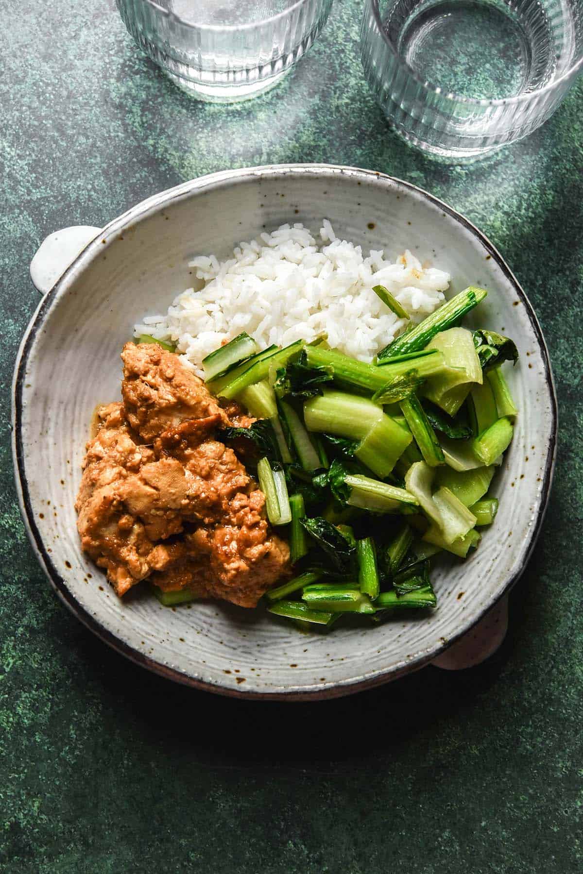 An aerial image of a bowl filled with marinated tofu, stir fried Asian greens and rice on an olive green backdrop