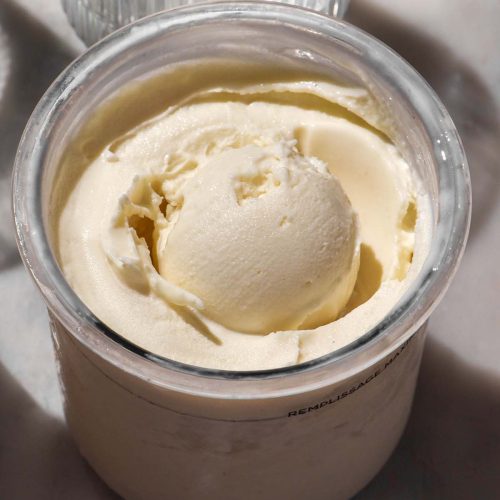 A brightly lit side on image of a tub of Ninja Creami frozen yoghurt on a white marble table. A sunlit glass of water sits in the back of the image.
