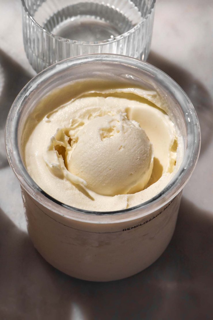 A brightly lit side on image of a tub of Ninja Creami frozen yoghurt on a white marble table. A sunlit glass of water sits in the back of the image.
