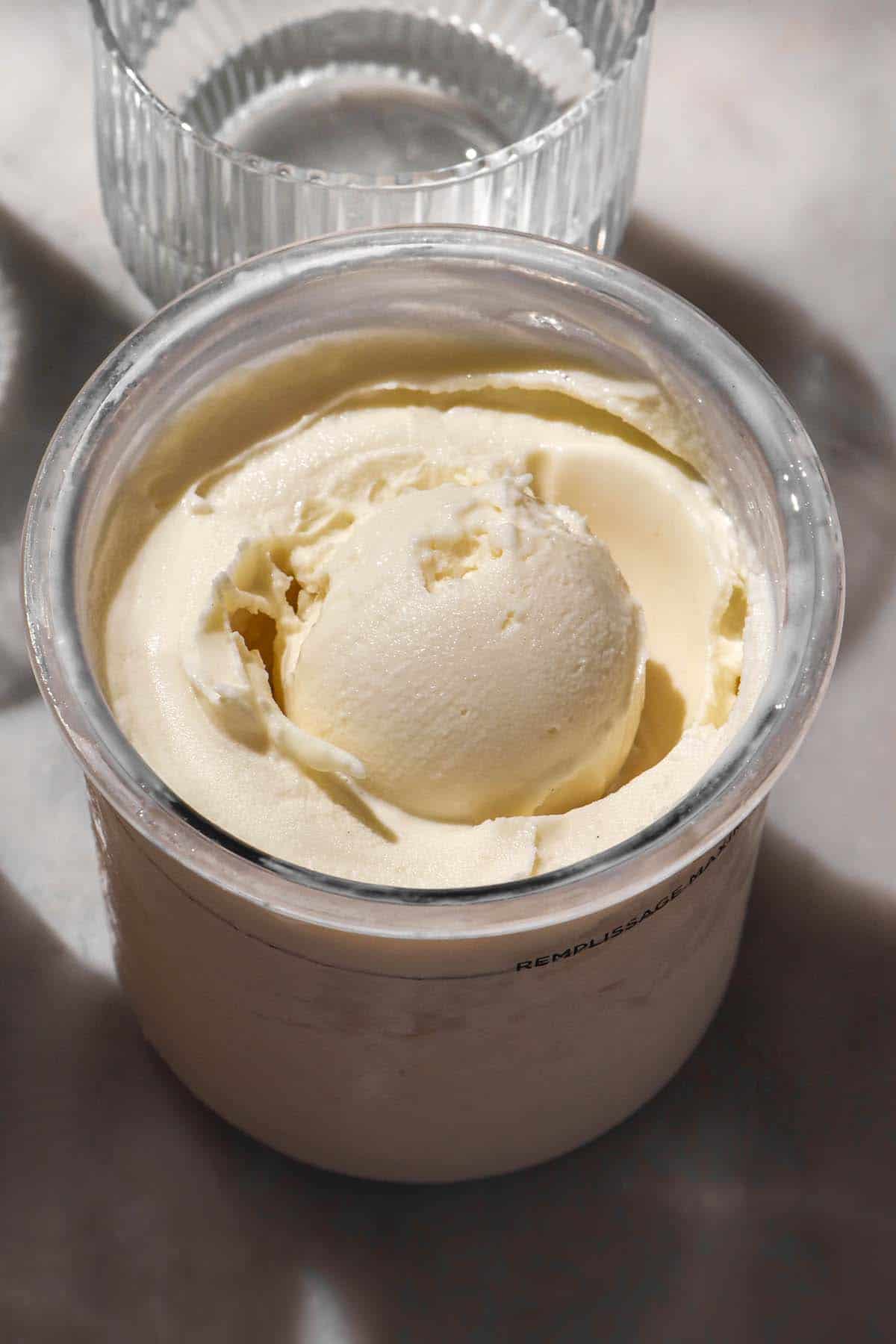 A brightly lit side on image of a tub of Ninja Creami frozen yoghurt on a white marble table. A sunlit glass of water sits in the back of the image.