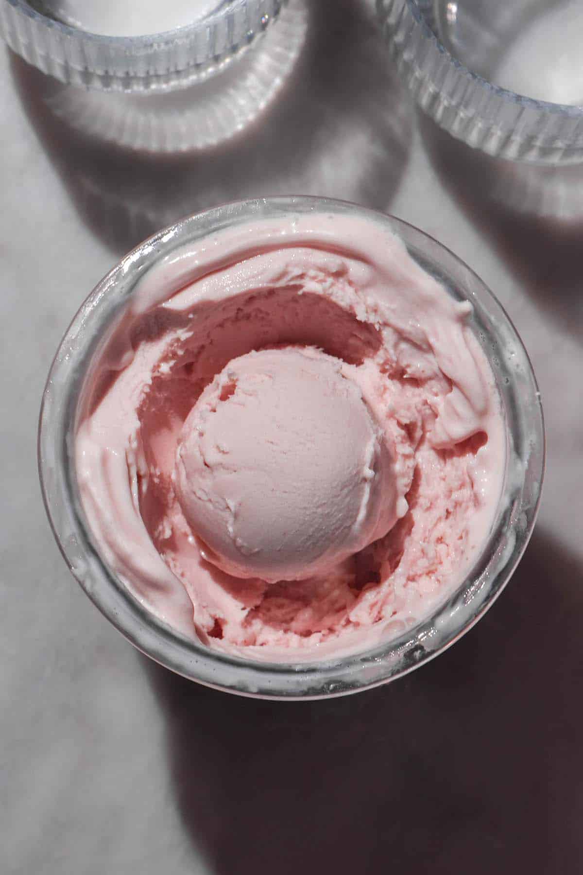 An aerial image of a tub of Ninja Creami vegan frozen yoghurt on a white marble table in bright sunlight. Two glasses of water sit in the top of the image.