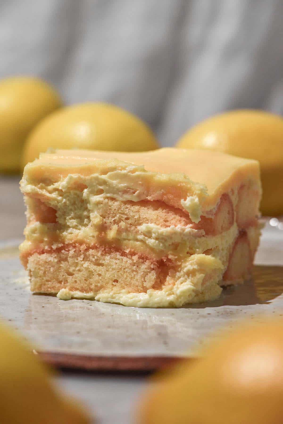 A side on macro image of a slice of gluten free Yuzu Tiramisu. The slice sits atop a white ceramic plate against a white linen backdrop. It is surrounded by lemons in the foreground and background of the image.