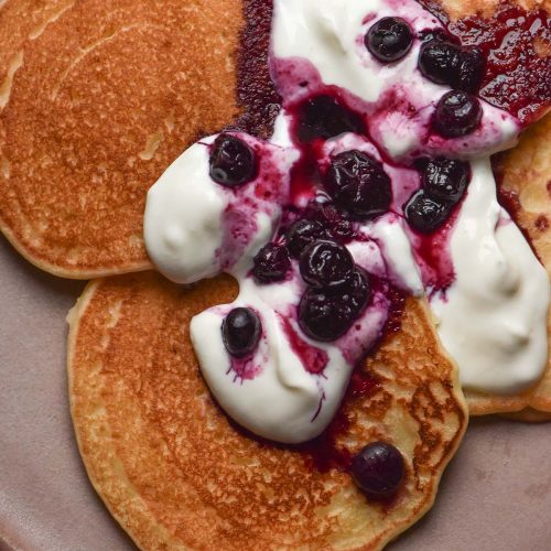 An aerial close up image of gluten free dairy free pancakes on a pale pink ceramic plate. The pancakes are topped with yoghurt and a blueberry coulis.