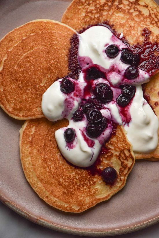An aerial close up image of gluten free dairy free pancakes on a pale pink ceramic plate. The pancakes are topped with yoghurt and a blueberry coulis.
