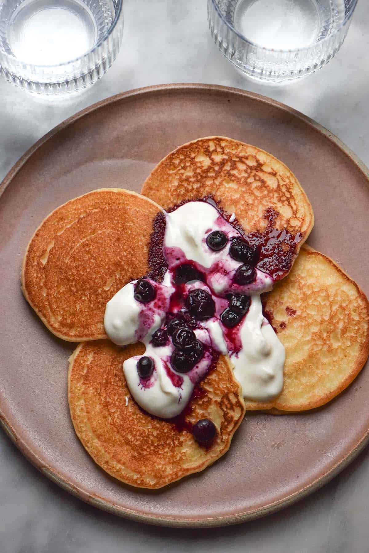 An aerial image of a pale pink ceramic plate topped with gluten free dairy free pancakes on a white marble table. The pancakes have been topped with yoghurt and a blueberry coulis.