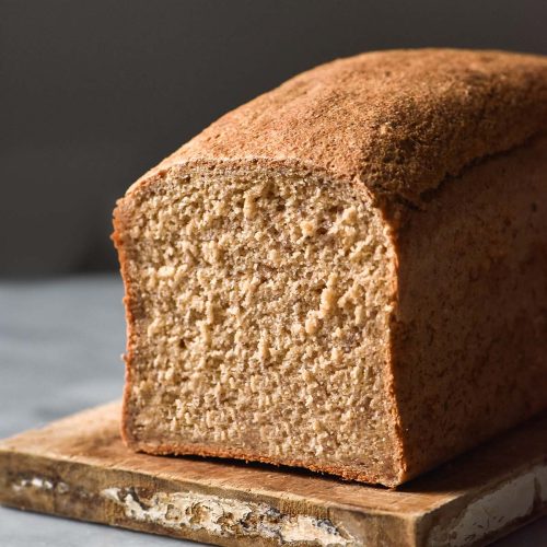 A side on view of a loaf of gluten free oat bread on a white marble table against a dark backdrop. The first slice of the oat bread has been removed, revealing the soft crumb.