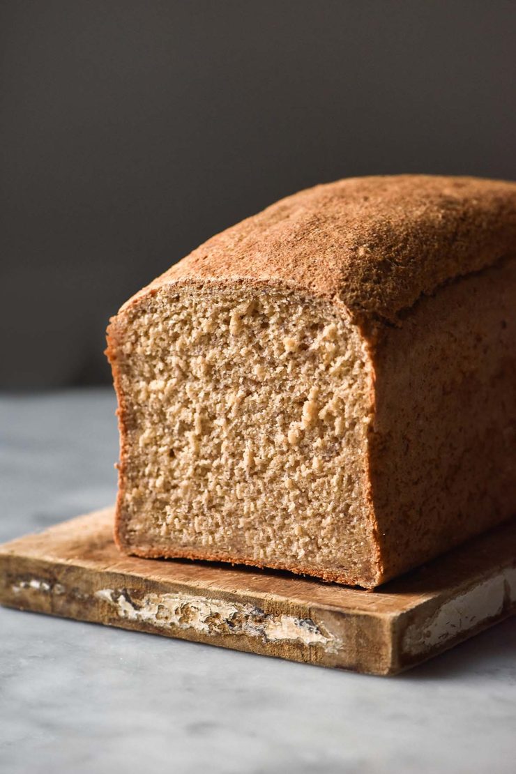 A side on view of a loaf of gluten free oat bread on a white marble table against a dark backdrop. The first slice of the oat bread has been removed, revealing the soft crumb.