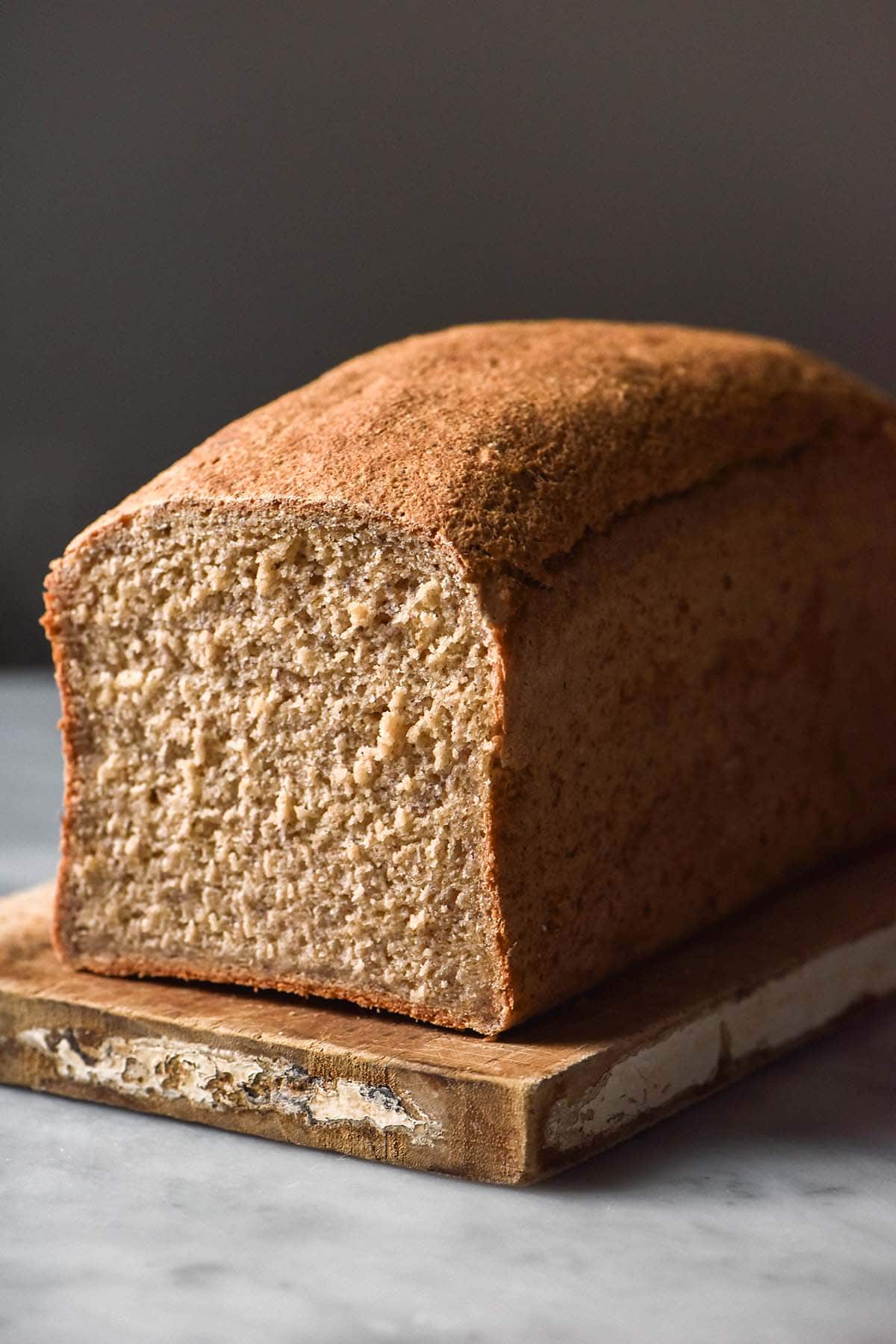 A side on image of a loaf of gluten free oat bread on a chopping board. The chopping board sits on a white marble table against a dark backdrop. The loaf has been sliced revealing the soft crumb inside. 