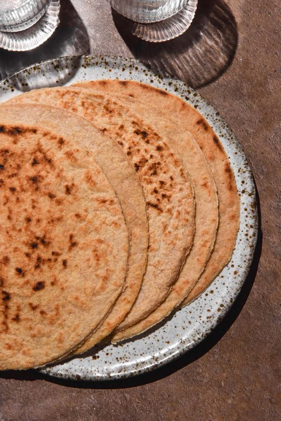 An aerial image of gluten free oat wraps fanned out on a white speckled ceramic plate atop a brown backdrop. Sunlit water glasses sit in the top left of the image.