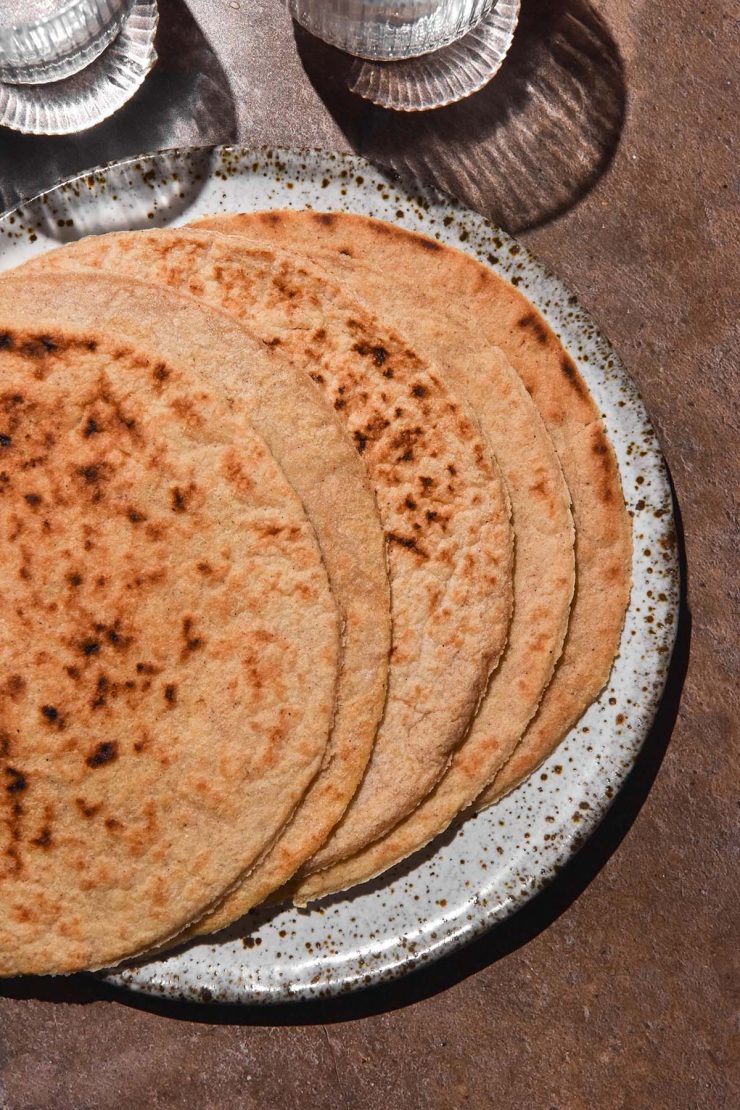 An aerial image of gluten free oat wraps fanned out on a white speckled ceramic plate atop a brown backdrop. Sunlit water glasses sit in the top left of the image.