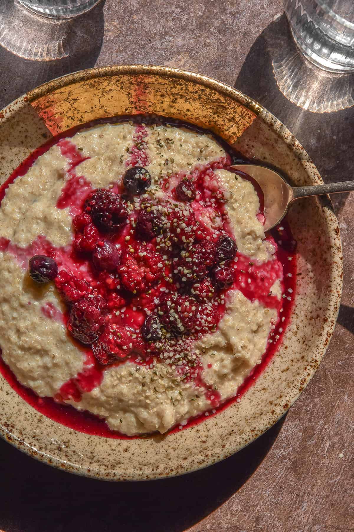 An aerial image of a bowl of quinoa porridge on a brown backdrop in bright sunlight. The porridge is topped with berry coulis and a drizzle of maple syrup