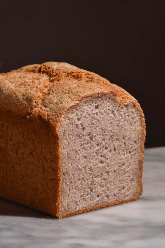 A side on view of a loaf of rice flour bread on a white marble table against a dark backdrop. The bread has been sliced to reveal a soft crumb inside.