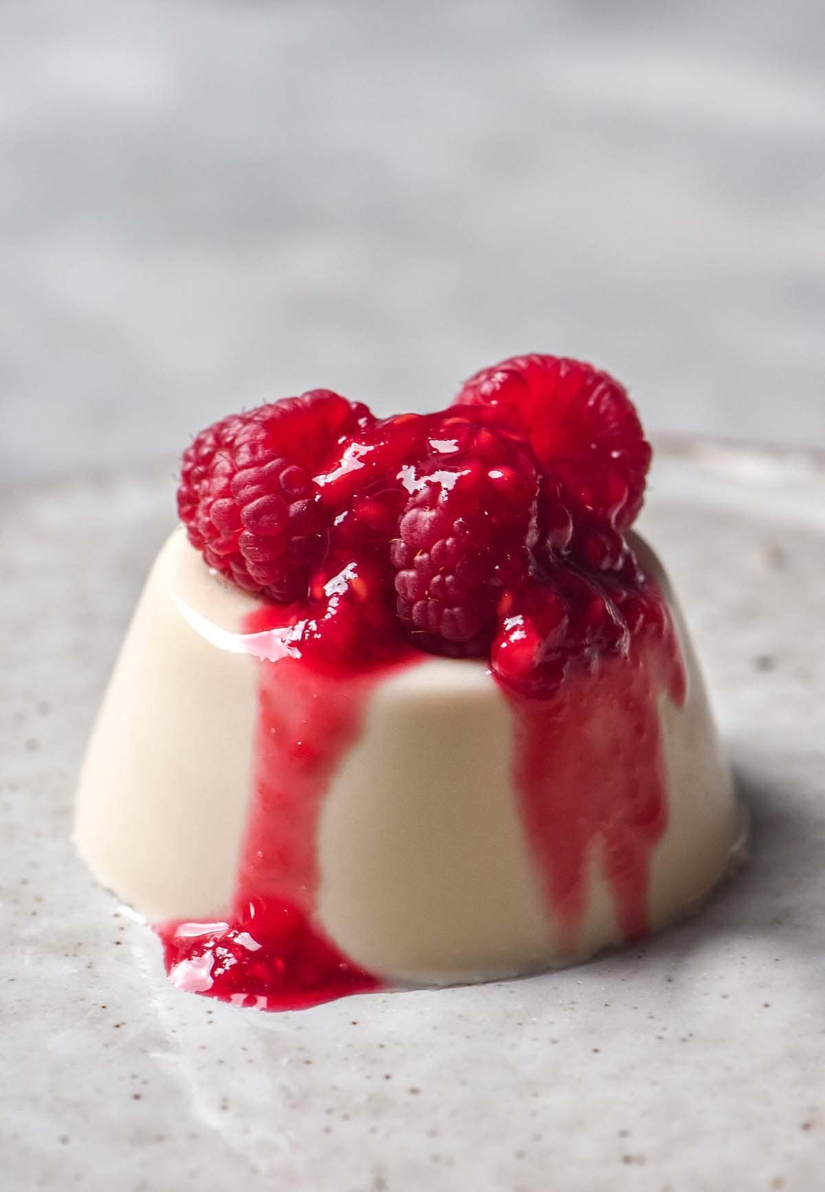 A side on image of a vegan panna cotta topped with raspberry coulis on a white ceramic plate atop a white marble table