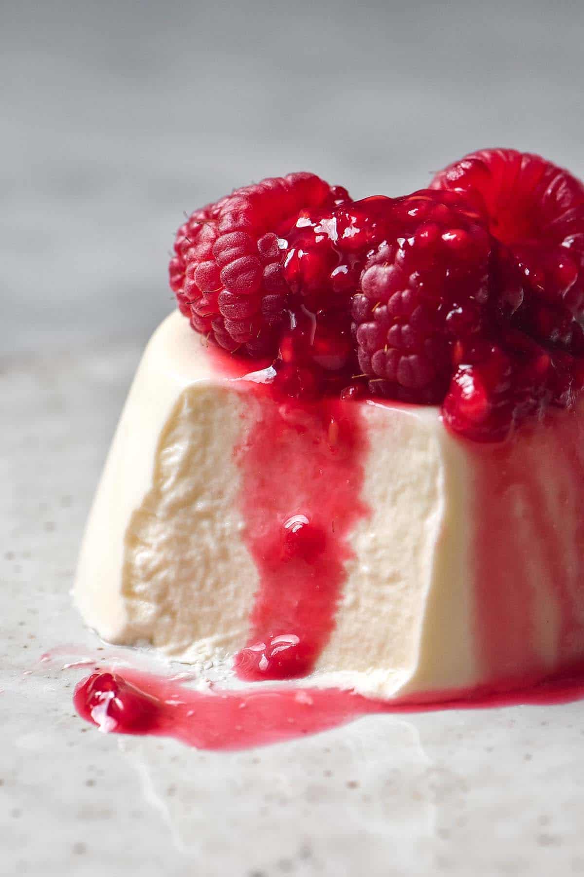 A side on image of a vegan panna cotta without coconut on a white ceramic plate atop a white marble table. The panna cotta is topped with fresh raspberries and raspberry coulis and a spoonful has been taken from the front left side of the panna cotta