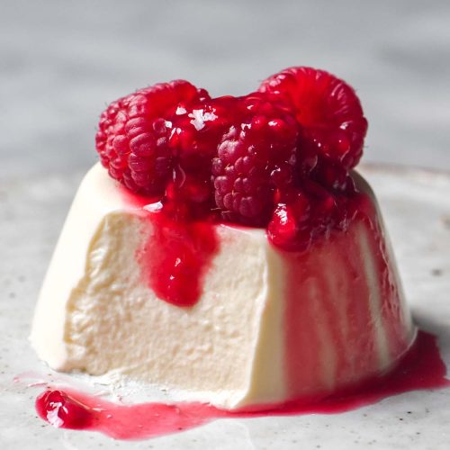 A side on image of a vegan panna cotta without coconut on a white ceramic plate atop a white marble table. The panna cotta is topped with fresh raspberries and raspberry coulis and a spoonful has been taken from the front left side of the panna cotta