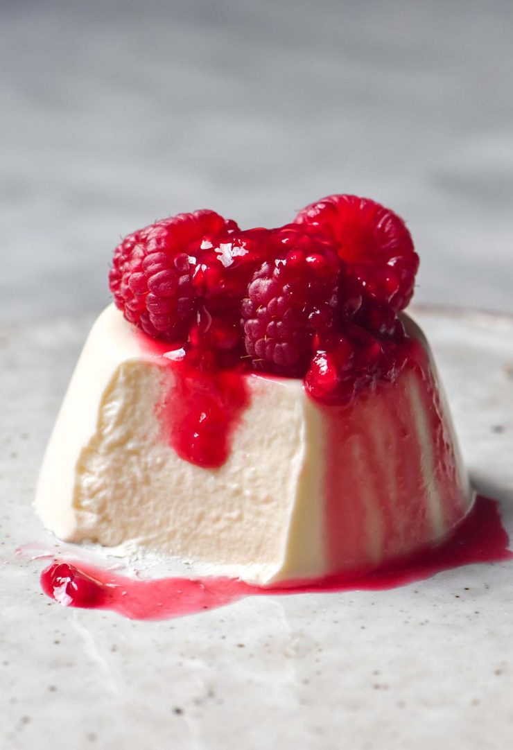 A side on image of a vegan panna cotta without coconut on a white ceramic plate atop a white marble table. The panna cotta is topped with fresh raspberries and raspberry coulis and a spoonful has been taken from the front left side of the panna cotta