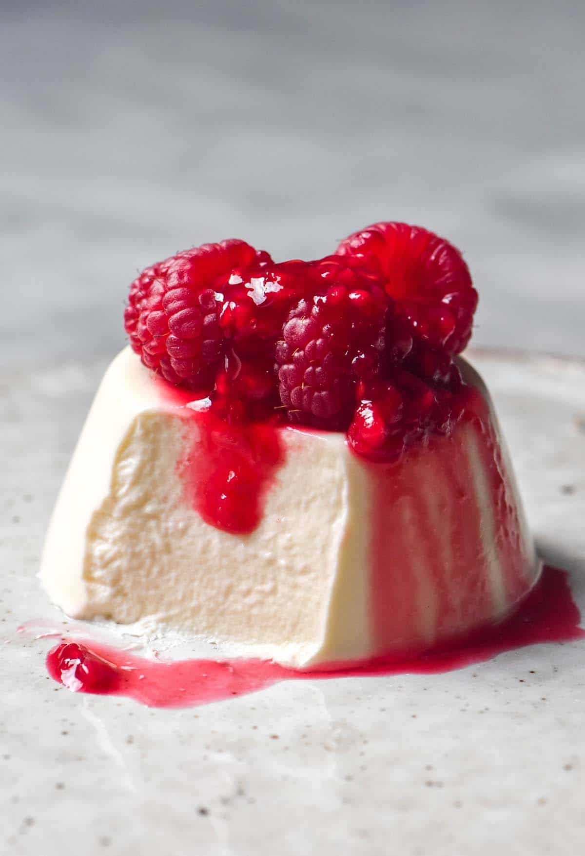 A side on image of a vegan panna cotta without coconut on a white ceramic plate atop a white marble table. The panna cotta is topped with fresh raspberries and raspberry coulis and a spoonful has been taken from the front left side of the panna cotta