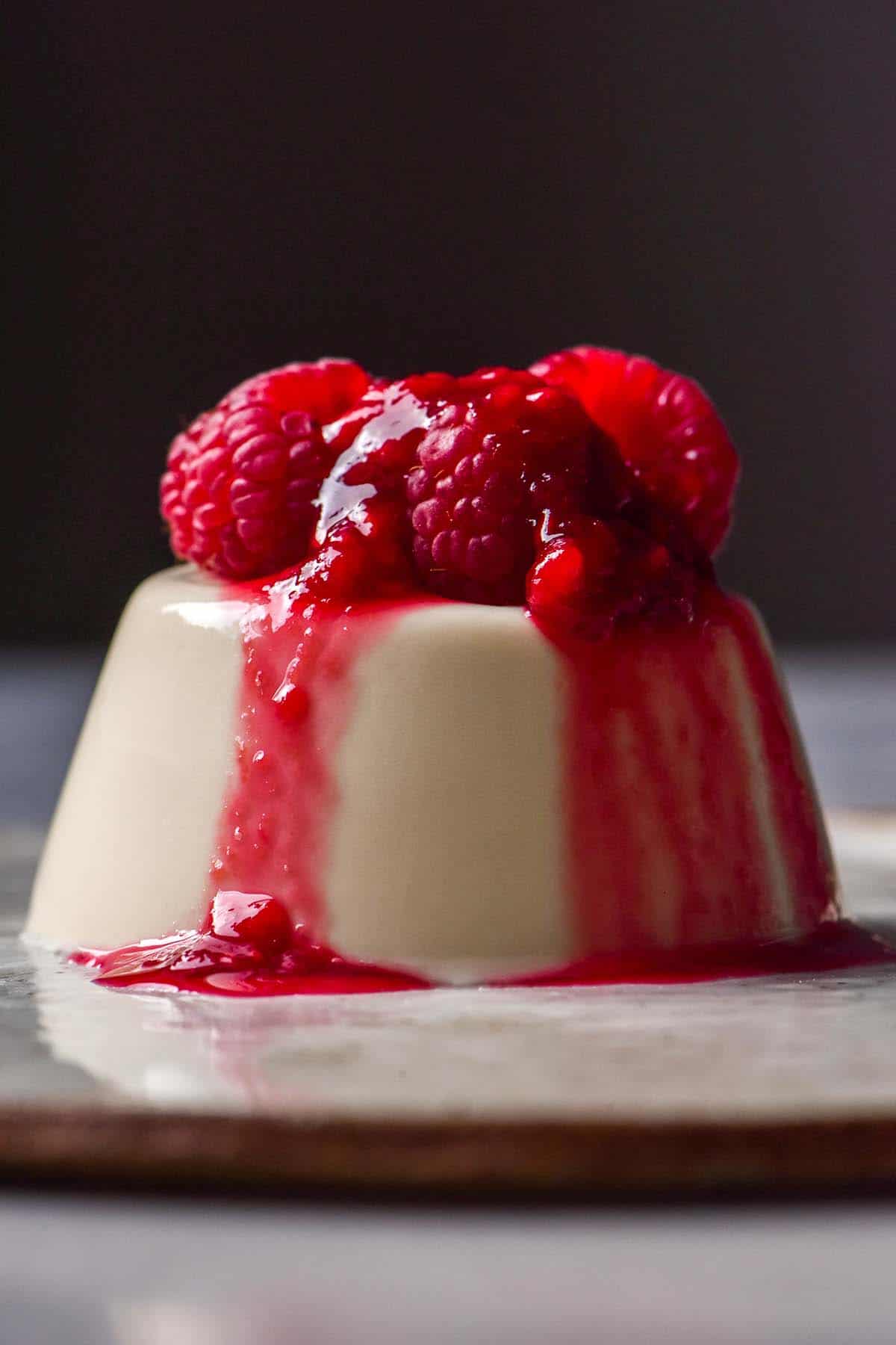A side on image of a vegan panna cotta topped with raspberry coulis. The panna cotta sits atop a white ceramic plate against a dark backdrop