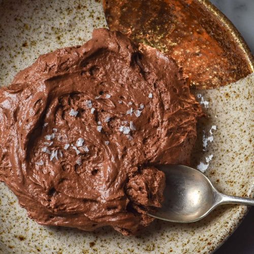 An aerial image of a beige ceramic speckled bowl filled with 3 ingredient chocolate mousse. The bowl sits on a white marble table and a glass of water sits in the top right corner.